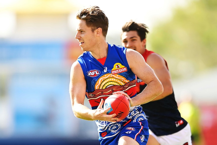 A Western Bulldogs AFL player runs with the ball in two hands as he his chased by a Melbourne opponent.