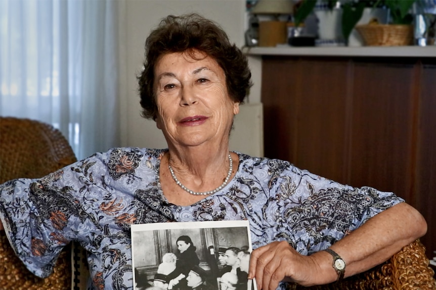 An older woman sits in an armchair holding a black and white photo.