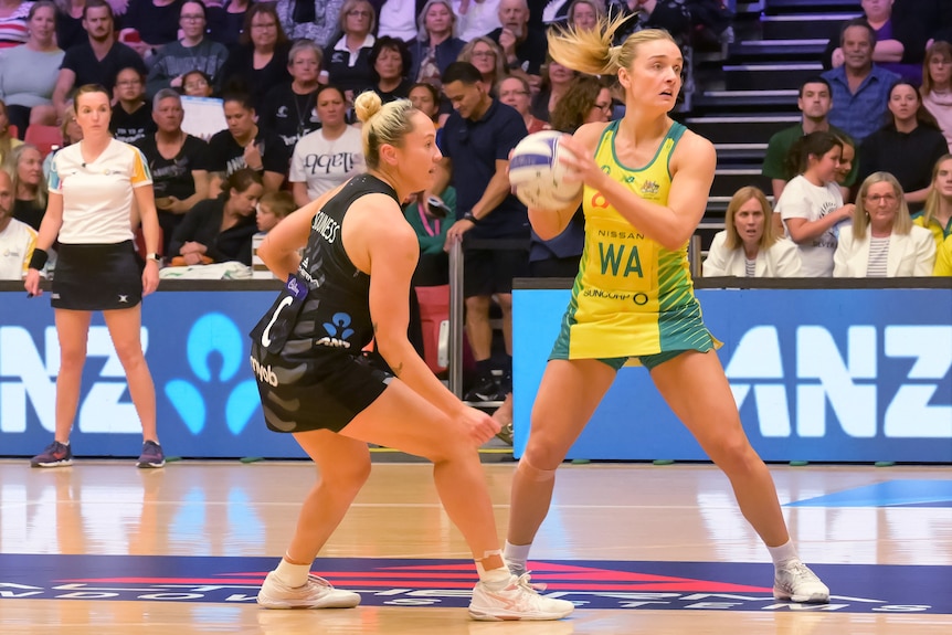 An Australian netballer holds the ball while under defensive pressure from a New Zealand opponent.
