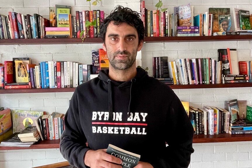 A man in a black jump which reads 'Byron Bay Basketball' standing in front of a bookshelf.