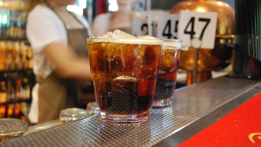 Two bourbon and coke's in close up on a bar, a female staff member stands out of focus in the background.