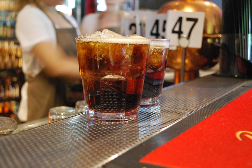 Two bourbon and coke's in close up on a bar, a female staff member stands out of focus in the background.