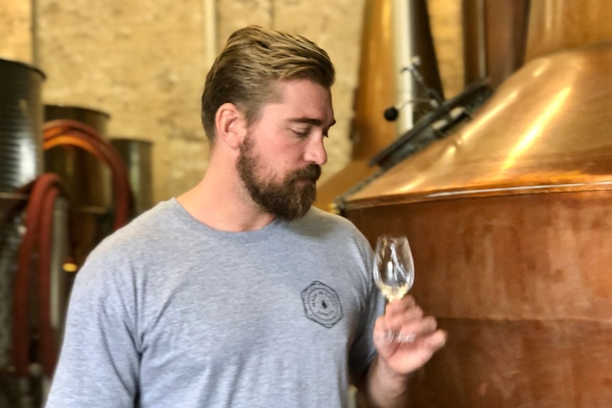 Trynt Xavier holds a glass, standing in front of copper stills.