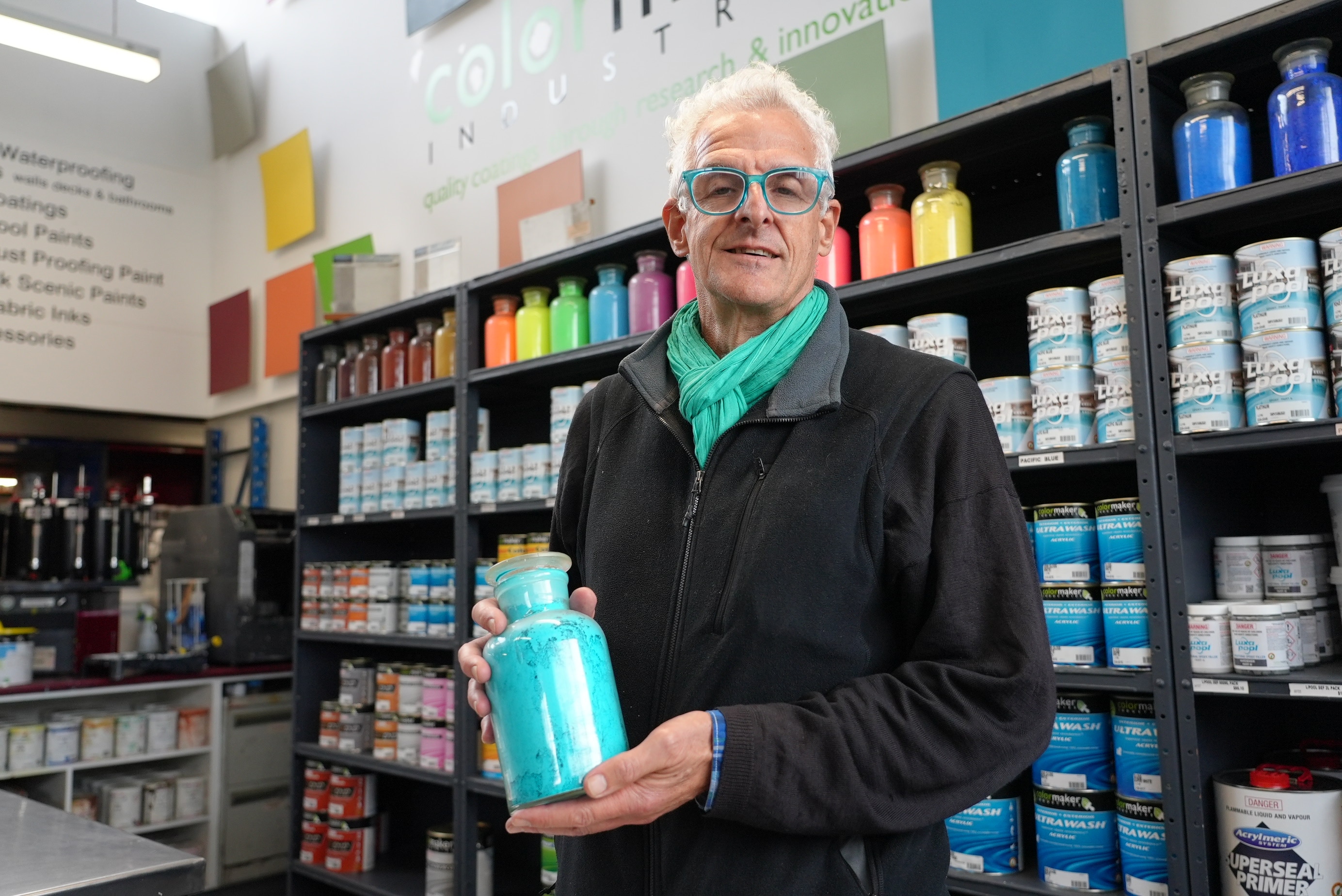 Man with grey hair and bright blue glasses stands in front of shelves stacked with brightly coloured paints.