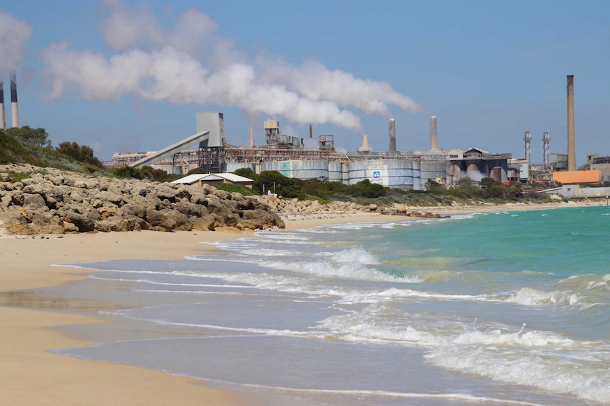 An empty beach leads to a heavy industrial area with large smoke plumes