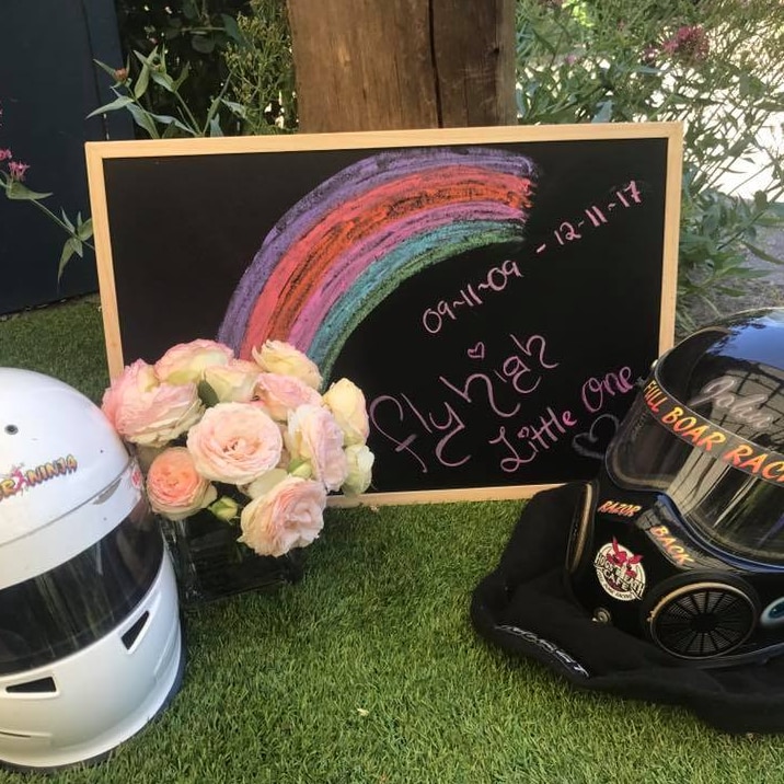 Two racing helmets sit outside on grass next to a bunch of flowers and a blackboard reading 'Fly high little one'.