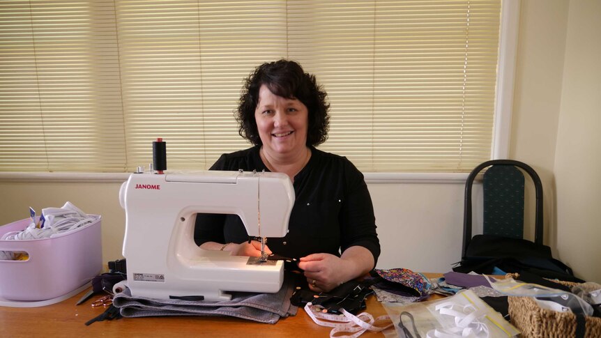 A woman with dark hair sits in front of her sewing machine and some face masks she's made