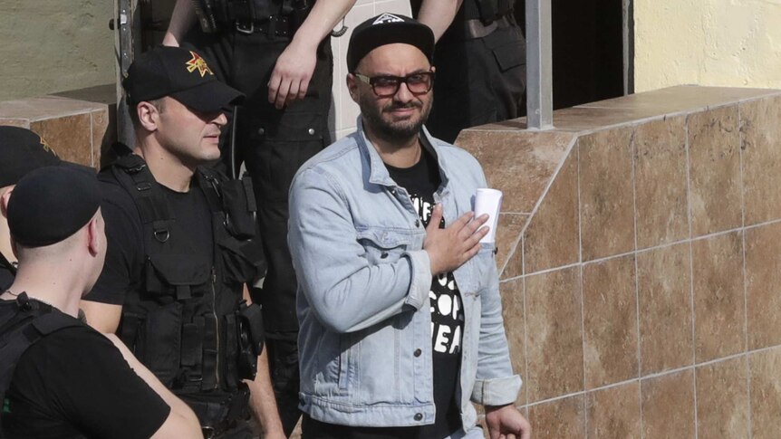 A group of police officers surround a man in a denim jacket, baseball hat and sunglasses.