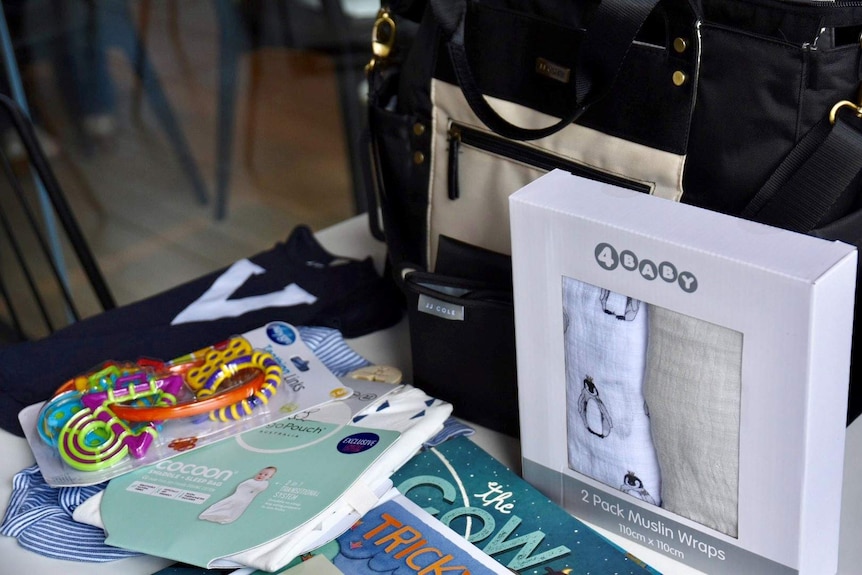 Items including baby clothes, a nappy bag, sleeping bag, muslin wraps, a teething toy and children's books are shown on a table