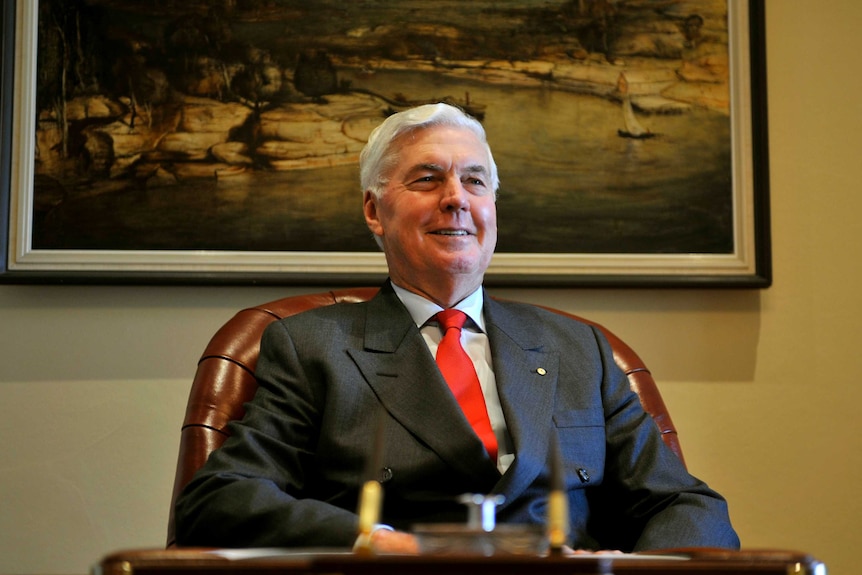 Michael Jeffery sits at a desk wearing a suit and red tie with a painting on the wall behind him.