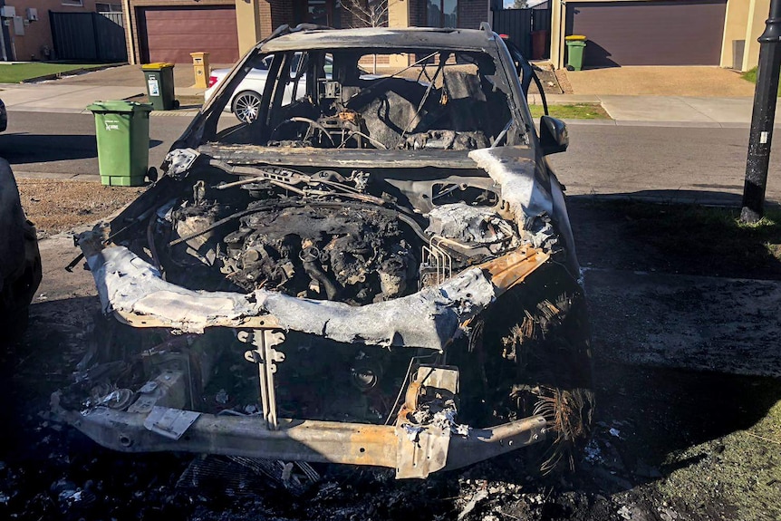 A burnt out Mercedes chassis sits in a driveway, with suburban homes behind.