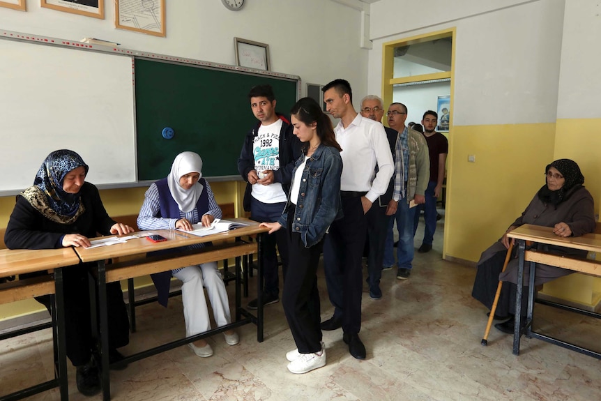 People queue to vote in a classroom. Two women sitting at a desk at the front of the classroom check voters' ids.
