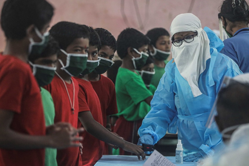 A string of Indian children wearing face masks are tested by a woman in blue protective gear.