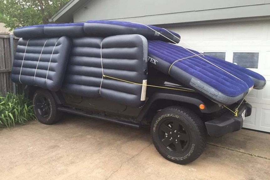 Air mattresses are tied on to the sides and roof of a four-wheel-drive car in front of a garage.