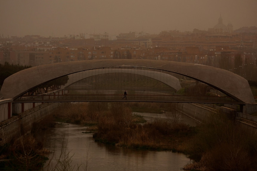 A person walks over a bridge with the sky tinted an orange-grey from dust.