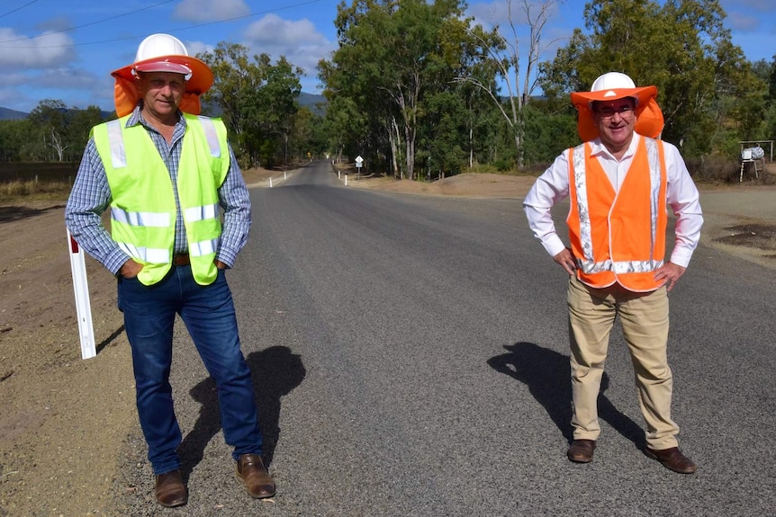 Two men in high visibility vest and hard hats, stand with hands on hips on newly completed bitumen road.