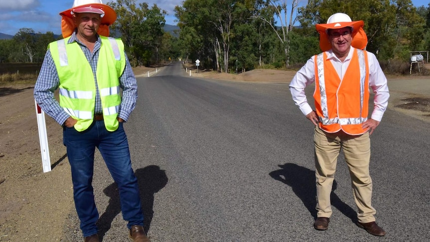 Two men in high visibility vest and hard hats, stand with hands on hips on newly completed bitumen road.