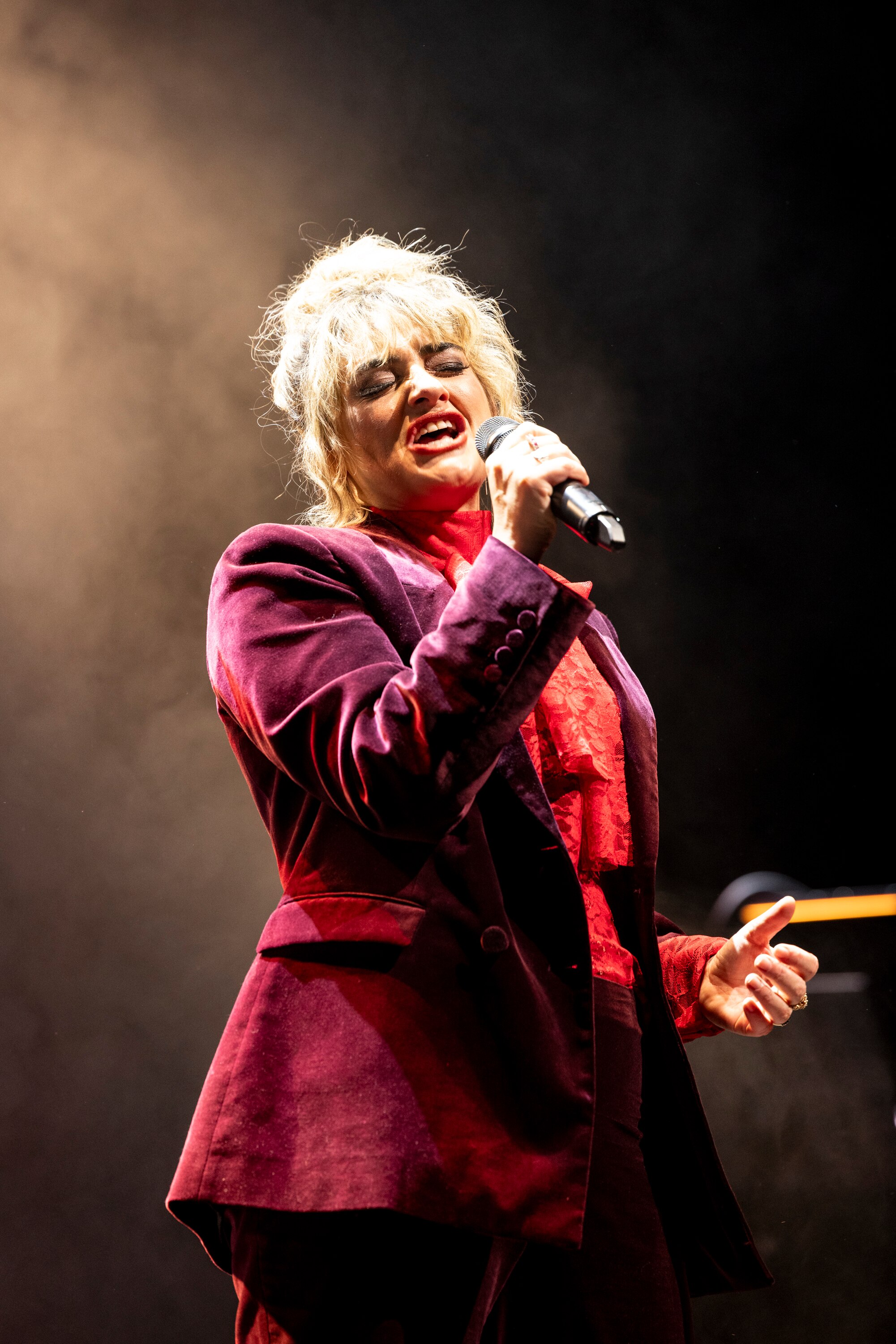 Person holds microphone and sings into with very focused expression, on smoky stage.