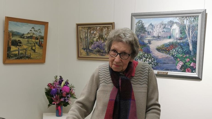 An elderly woman stands in front of several beautiful landscape paintings.