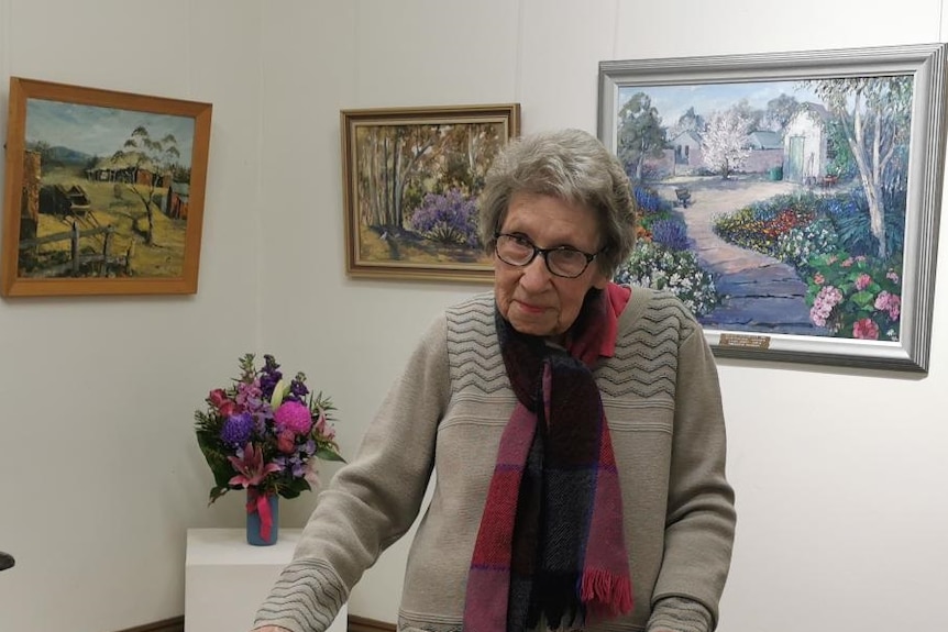An elderly woman stands in front of several beautiful landscape paintings.