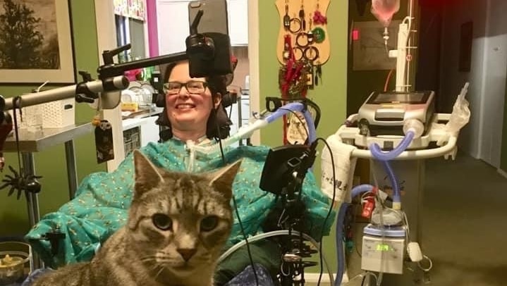 A woman lies on a medical bed surrounded by equipment and a cat at her feet