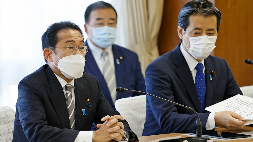 Two Japanese men in suits and face masks sit at table with mics in front, while a third man watches behind