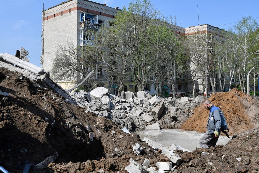 Man stands next to crater from explosion outside damaged apartment.