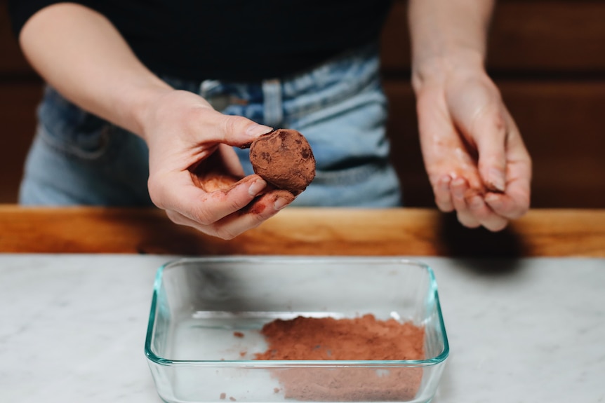 A person's hand holds a cocoa-dusted ball of chocolate above a glass dish filled with more cocoa.