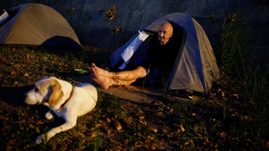 A man who is homeless, sits in a tent with his dog, as night falls.