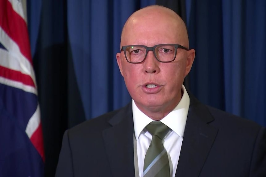 A white man with a bald head and glasses, wearing a suit, in front of a blue curtain with an Australian flag beside him.