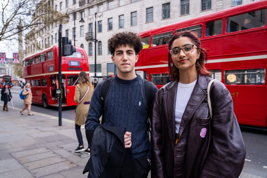 A young woman in a leather jacket and a young man in a blue jumper stand on a footpath in front of a london double decker bus