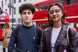 A young woman in a leather jacket and a young man in a blue jumper stand on a footpath in front of a london double decker bus