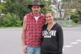 A man in a sleeveless flannel shirt and a hat stands on a suburban street with his arm around his wife.