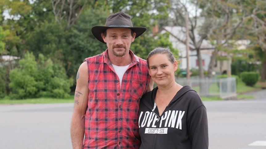 A man in a sleeveless flannel shirt and a hat stands on a suburban street with his arm around his wife.