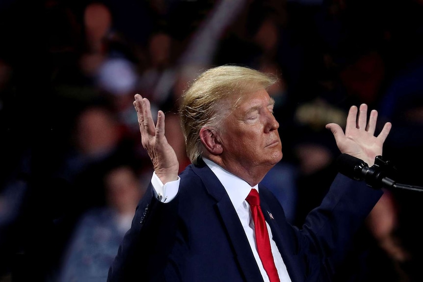 Donald Trump stands at a lecturn with his hands raised and his eyes closed