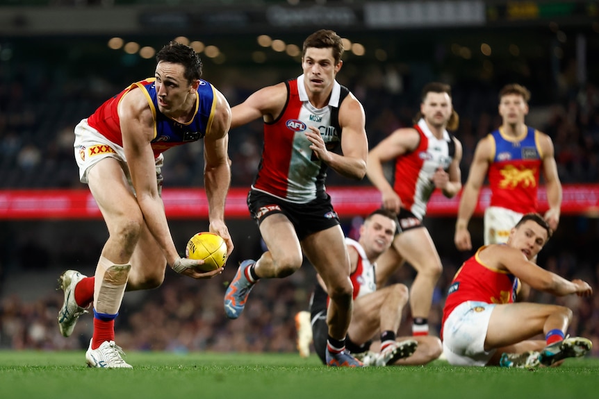 A Brisbane AFL ruckman bends down and scoops the ball ready to handball to a teammate as defenders watch.