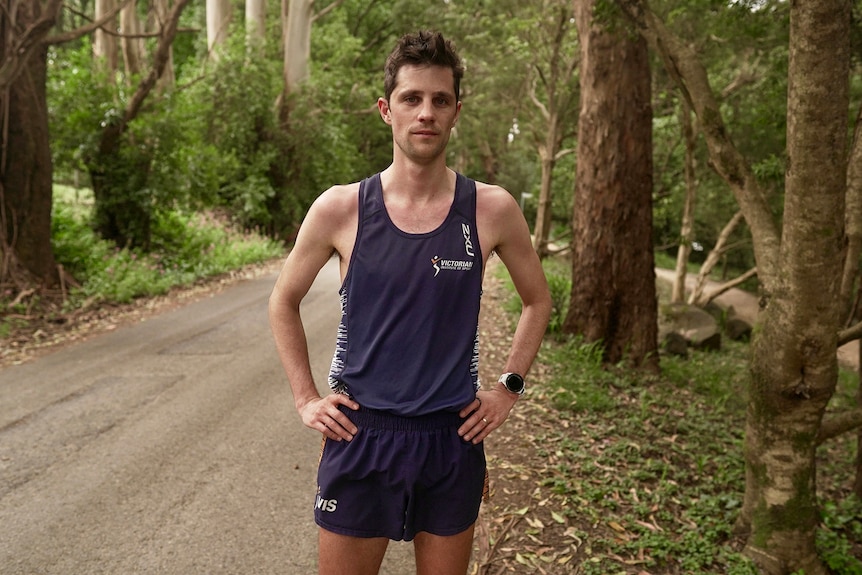 A man dressed in athletics wear poses for a photo on a track in a forest.