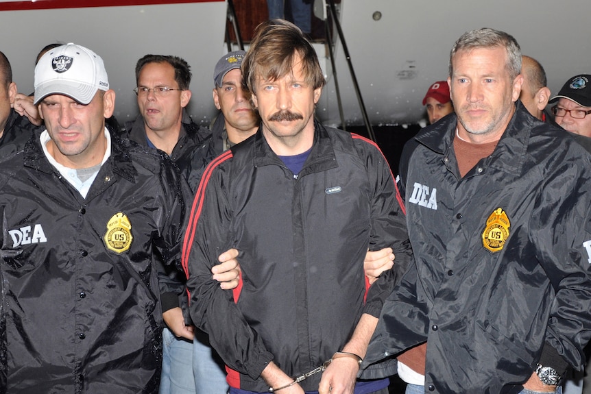A man with a moustache and handcuffs on is escorted off a plan by DEA officers