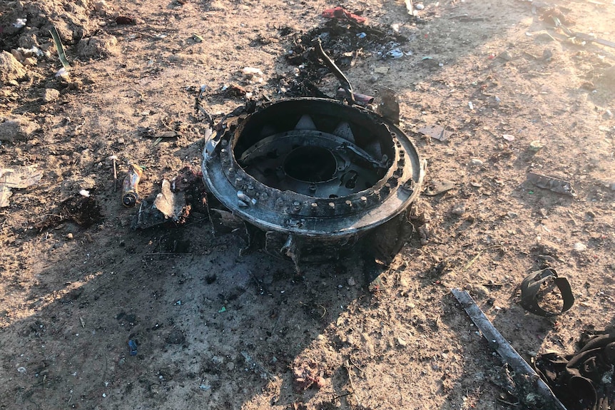 A round plane part is seen on the ground. It is surrounded by debris from the rest of the crash, and sits atop brown dirt.