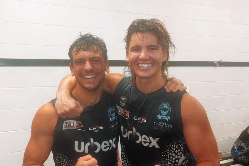 Two football players arm in arm smiling at the camera in the sheds