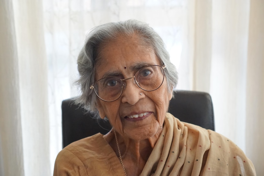 A woman with grey hair, thin-rimmed glasses and brown top and shawl smiles and looks ahead.