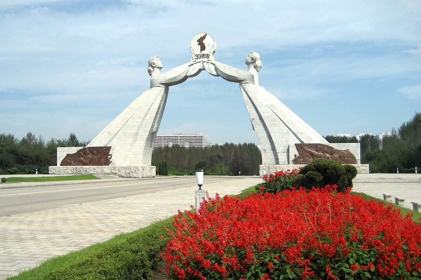 Stone statues of two women in hanboks joining hands to make an arch with red flowers in the foreground.