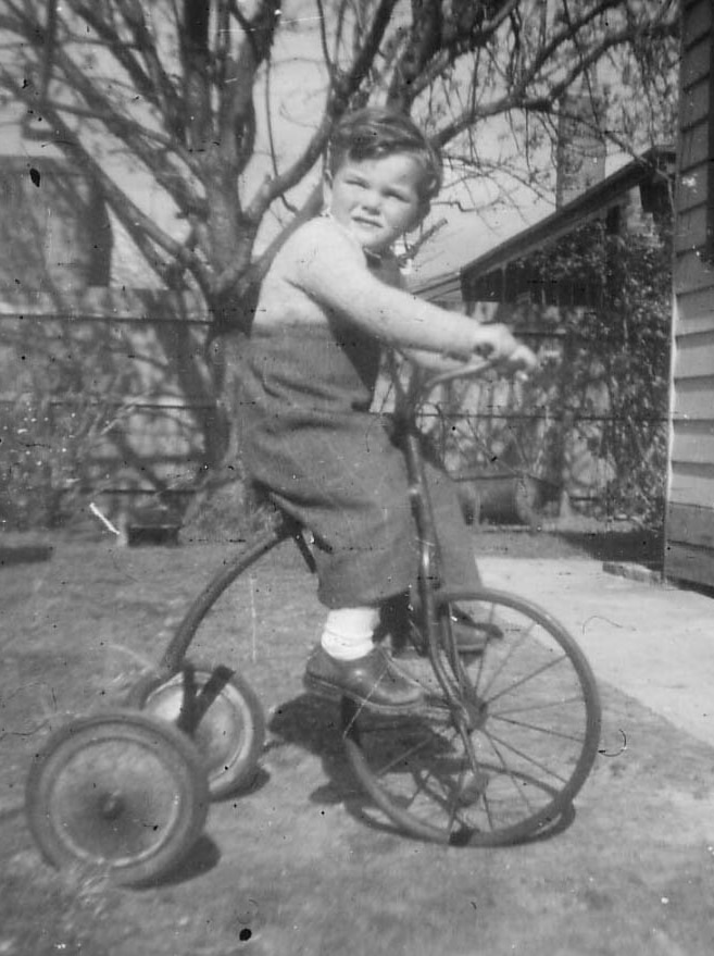 A black and white photo from 1956 showing a young boy riding a tricycle.