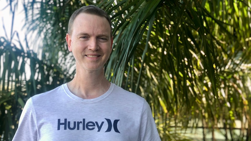 Man wearing t-shirt smiles at camera with palm trees in the background
