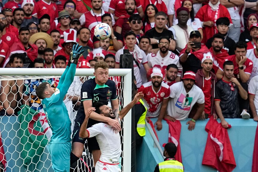 An Australian goalkeeper and defender fly for the ball in the box in front of a Tunisian forward.