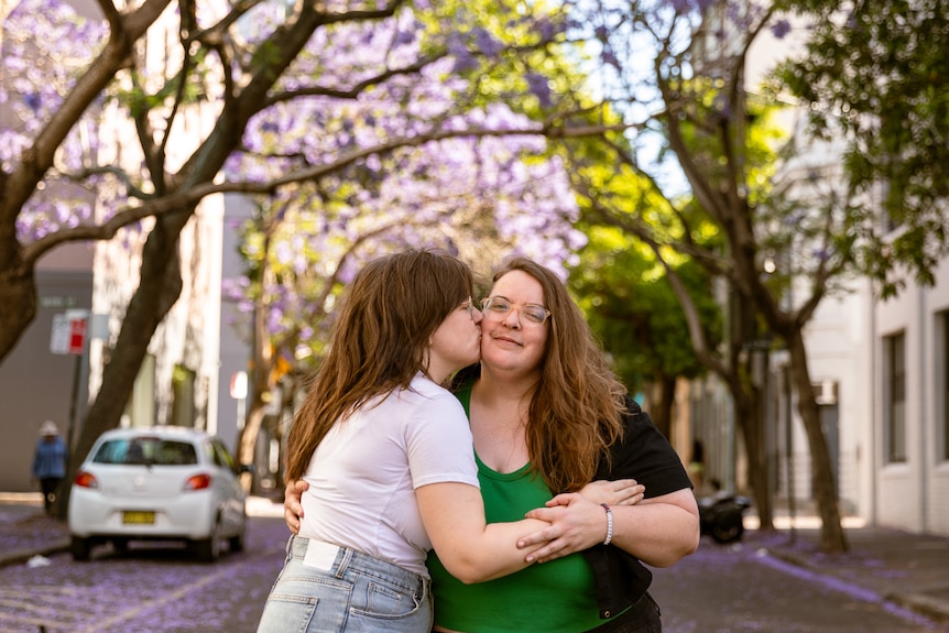 Freya, left, places and arm around Bec and plants a kiss on her cheek. Bec looks to the camera and smiles.