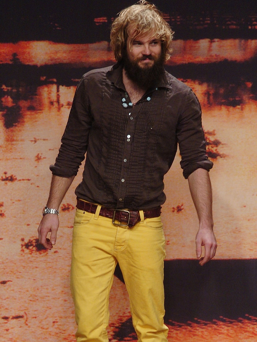 A bearded man with shaggy dark blonde hair stands at the end of a runway