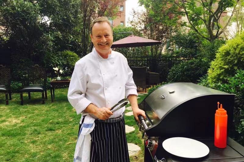 Australian expat Paul Tait holds tongs next to a barbecue grill smiling at a backyard.