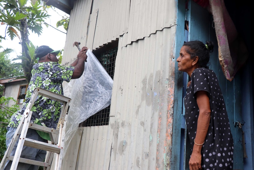 A man stands on a ladder and nails a sheet of plastic over the window of a home as a woman watches on