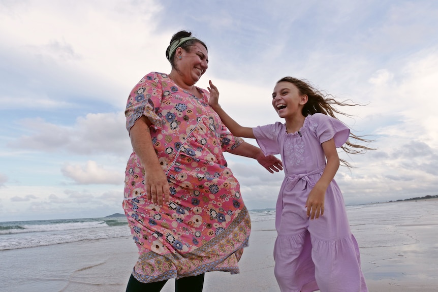 a woman in a pink flowy dress smiles at a young girl in a purple dress while standing on a beach